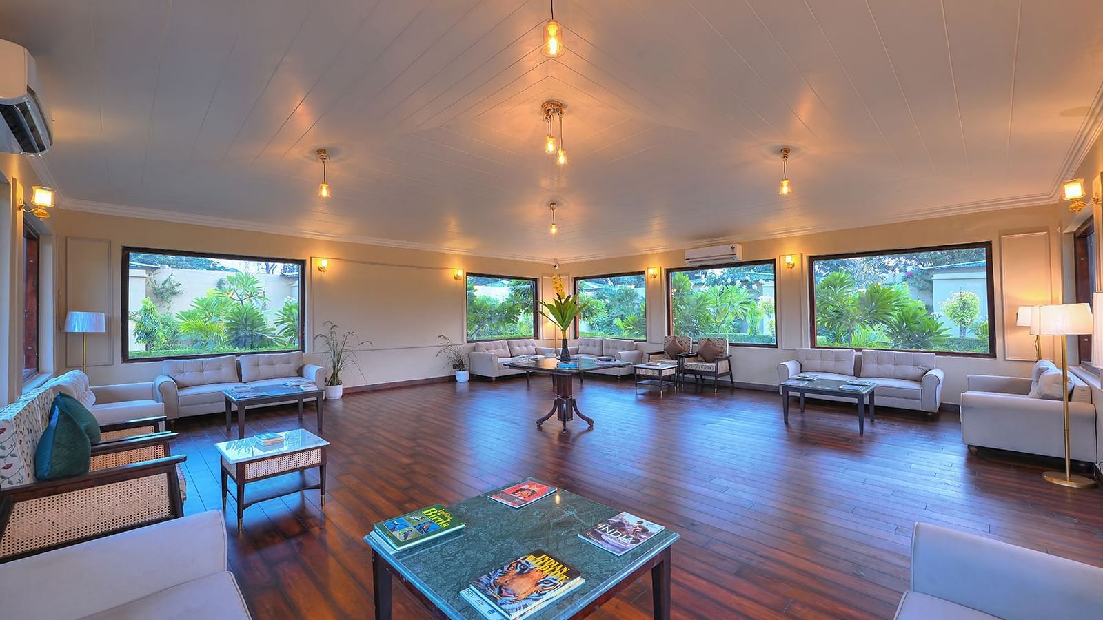 A spacious and well-lit lounge area featuring multiple seating arrangements, large windows overlooking greenery, and a polished wooden floor - Singhvilas, Ranthambore