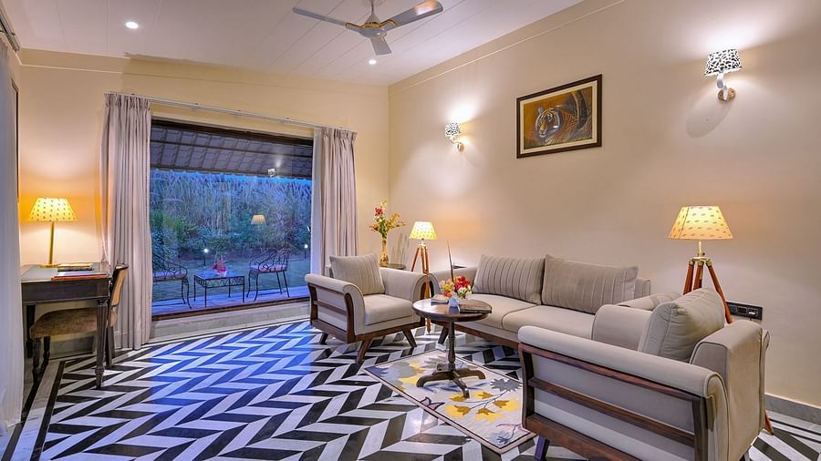 A cosy living room of Superior Cottage with beige sofas, a wooden table, patterned flooring, and a large window overlooking an outdoor seating area - Singhvilas, Ranthambore