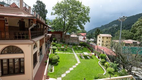 Aerial view of The Pavilion, Nainital's landscaped gardens with geometric lawn patterns, pathways, and traditional buildings nestled in forested hillside.