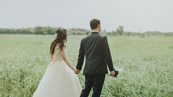 A bride and groom in wedding attires walking thorugh a field while holding hands