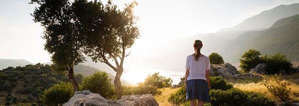 A person standing on a rock, next to a boulder, overlooking trees and hills under sunlight.