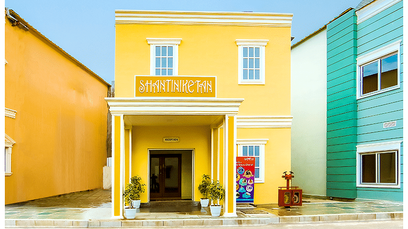 Front view of the bright yellow Shantiniketan building with white-framed windows, a reception entrance, and potted plants on either side.