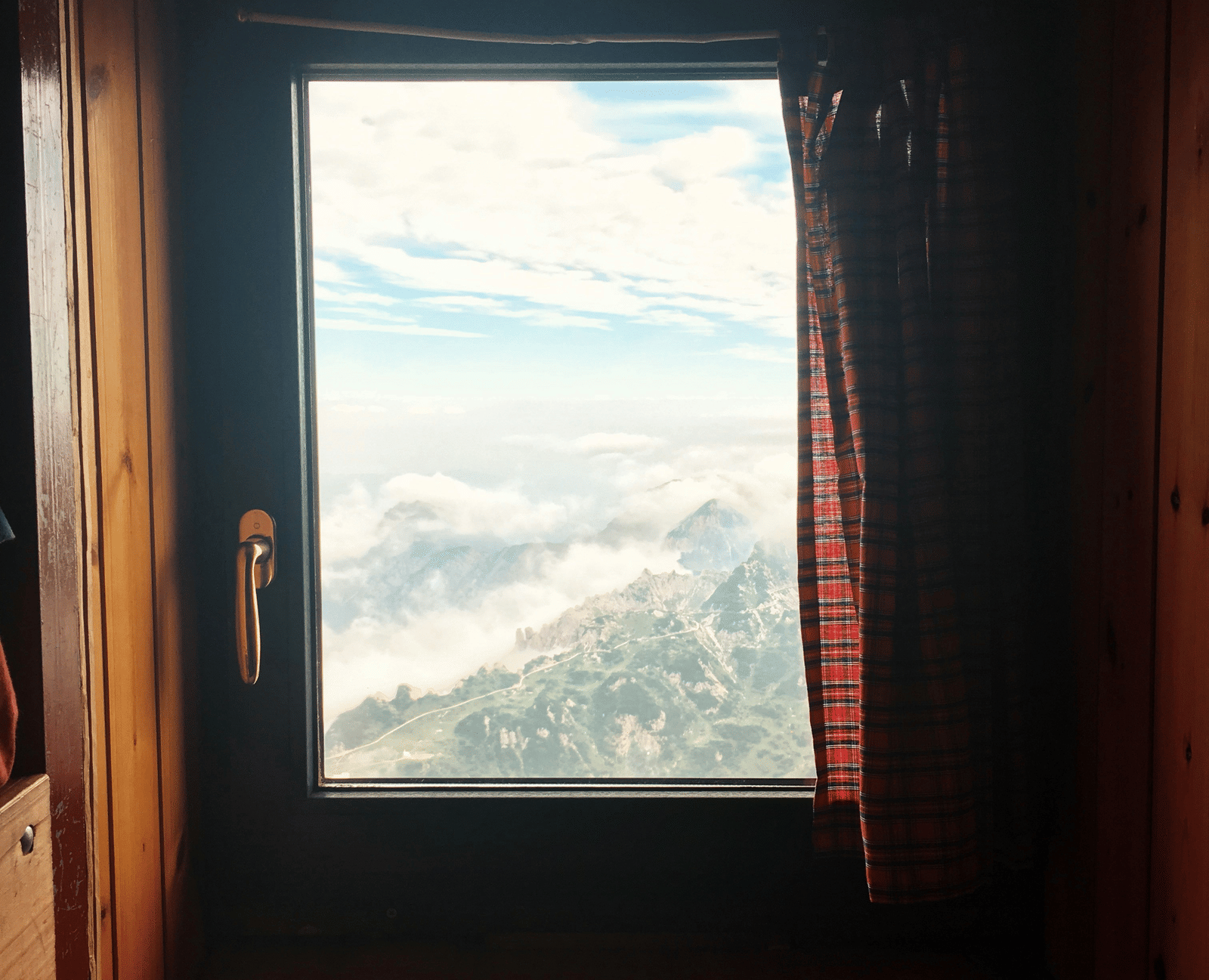 A window framed by dark wood, looking out onto a stunning vista of rolling, misty mountains under a bright, cloudy sky.