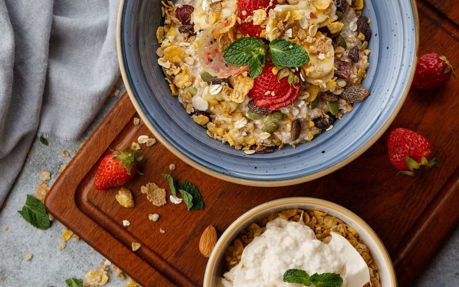 Top view of a breakfast bowl filled with granola, fresh strawberries, and mint leaves, accompanied by a smaller bowl with yoghurt and apple slices.