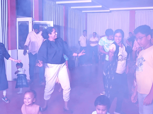 A blurry, vertical image showing a group of people, including some children, in a dimly lit indoor space with purple and white lighting, possibly at a lively event.