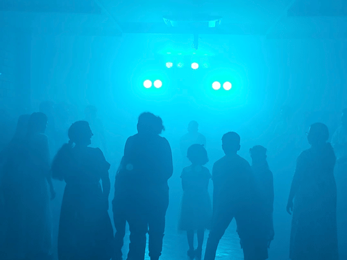 A vertical, dark image featuring silhouettes of several people standing in a blue-lit, foggy or smoky environment, creating an atmospheric and somewhat mysterious scene.
