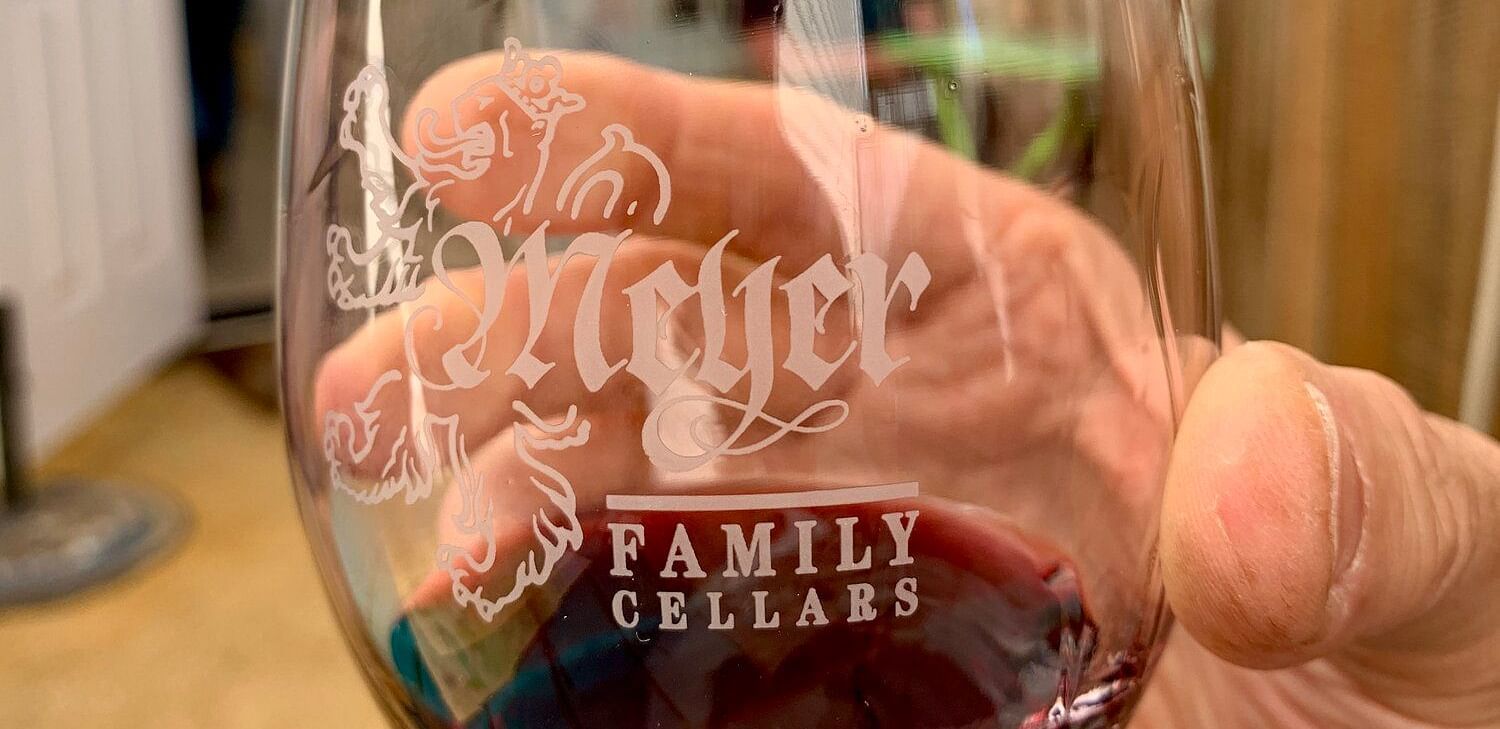 A hand holds a wine glass filled with red wine, featuring the Meyer Family Cellars logo.