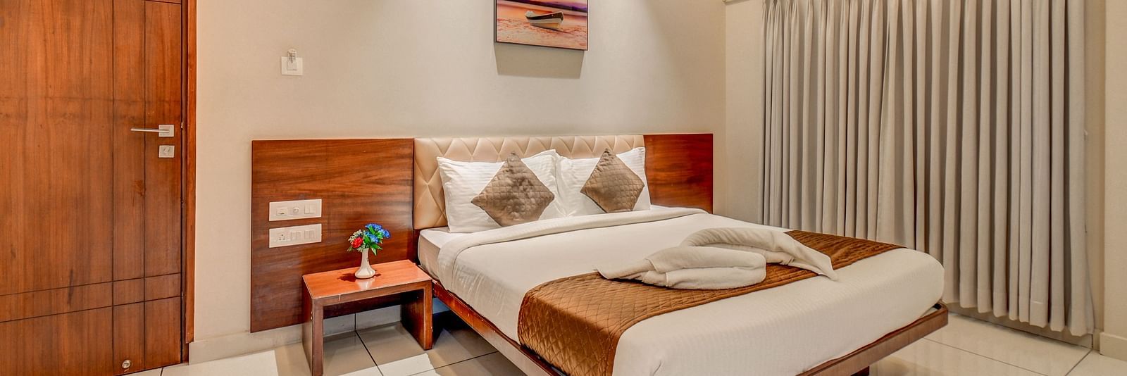 A spacious room at Harigovinda's JNStay with a large bed, wooden headboard, bedside table with a plant, a door, and curtains covering the window