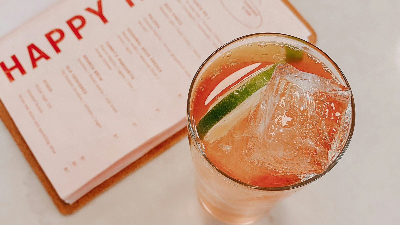 A top view of a drink with a wedge of lime placed next to a white card with Happy hour written in red letters