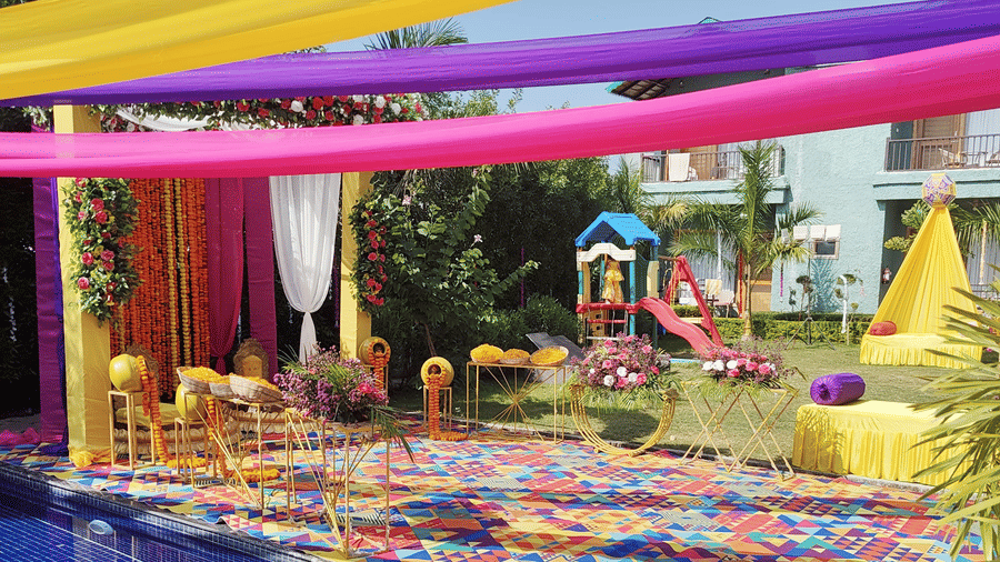 A view of the pool area decorated at Corbett Nirvana Resort with colourful yellow and pink fabric drapes overhead and patterned cushions near the water.