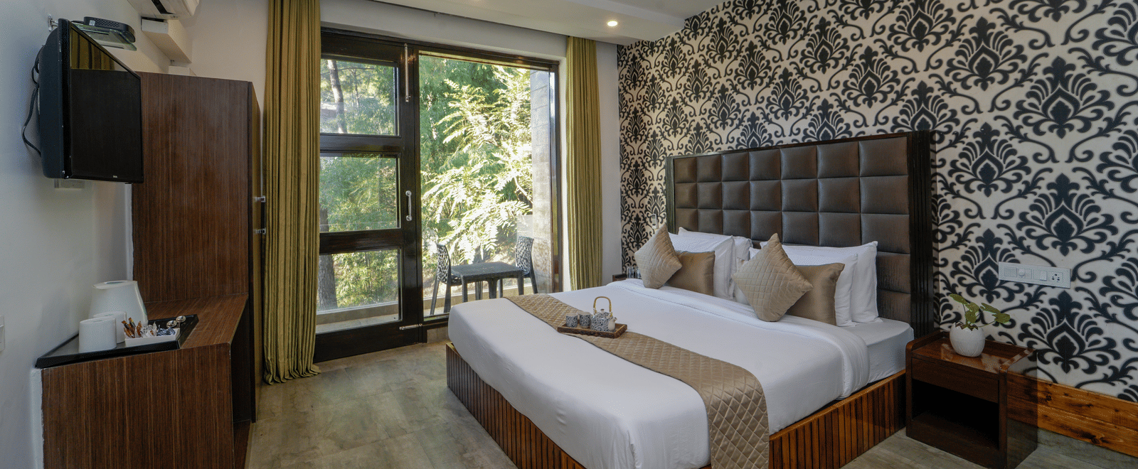 The deluxe room at DLS Whispering Winds, Kasauli, showcasing a bed, bedside tables, flat-screen TV, other room amenities, and a glass door leading to the balcony.