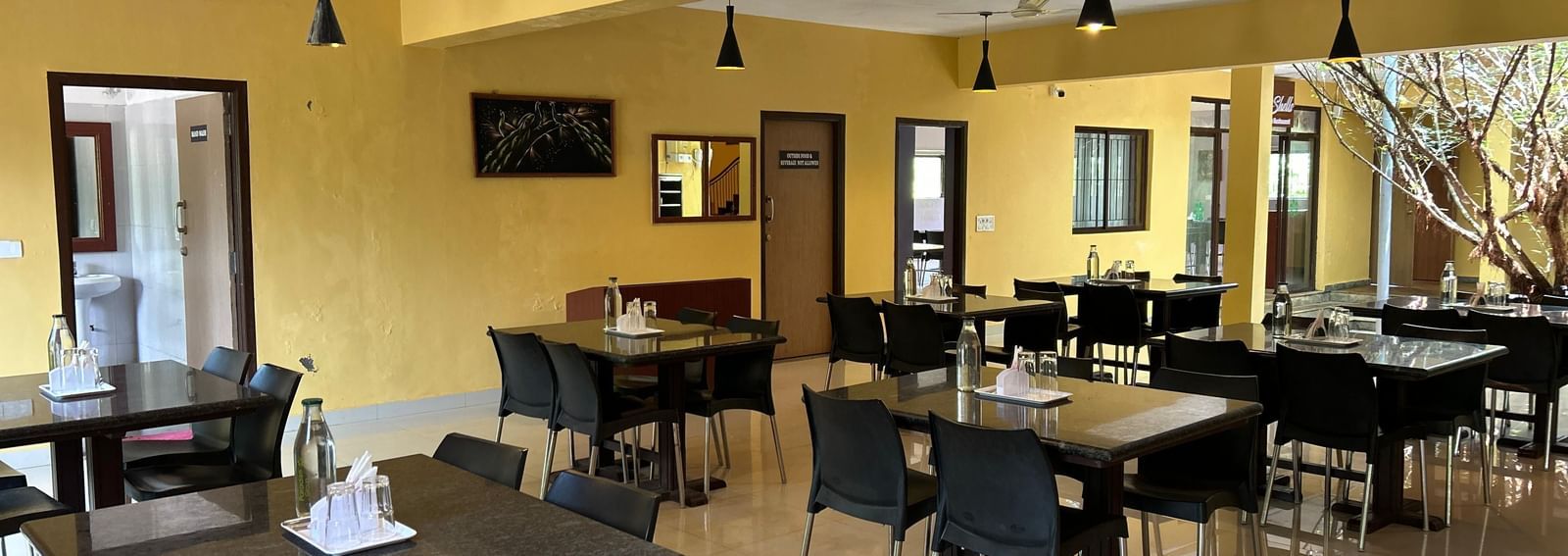 Dining room with yellow walls, tables, black chairs, and hanging lights at Paradise Wild Hills Resort, Kollur.