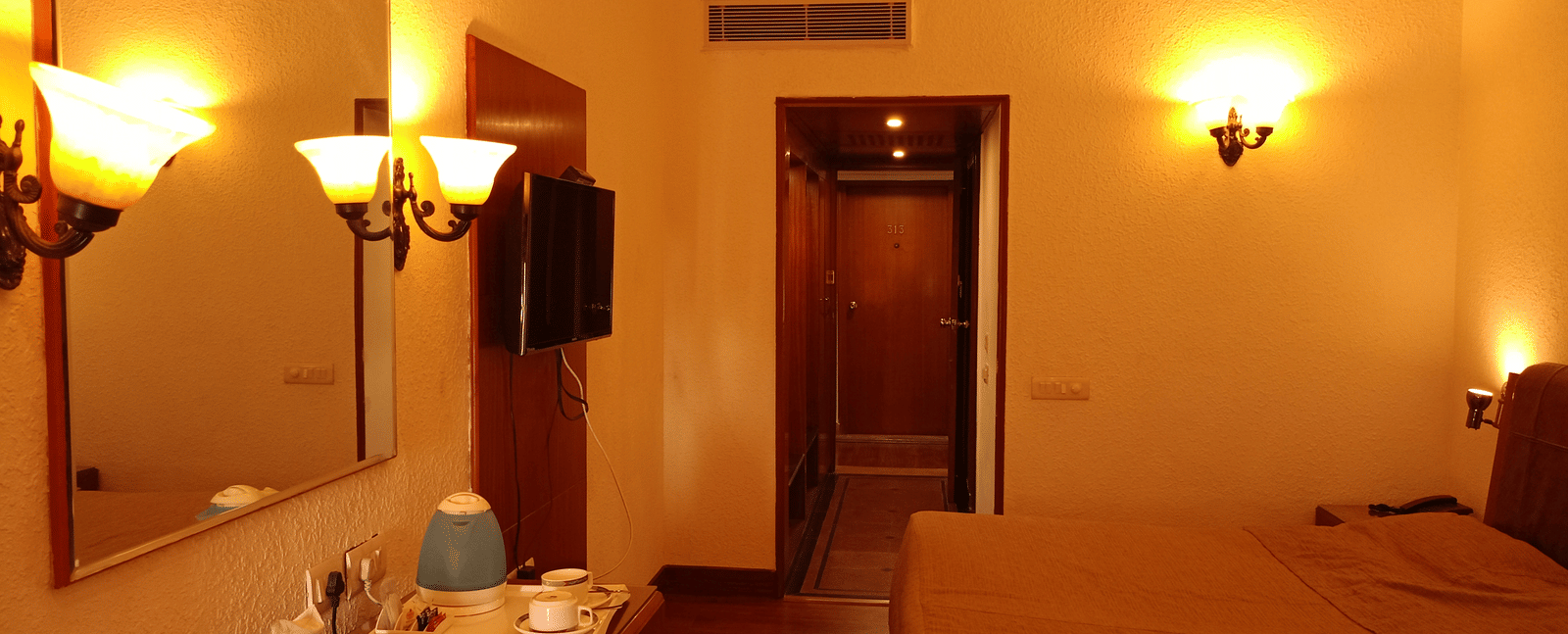 Room interior at Hotel Gold, Panipat with a bed, lamps, desk, and a mirror near the entrance.