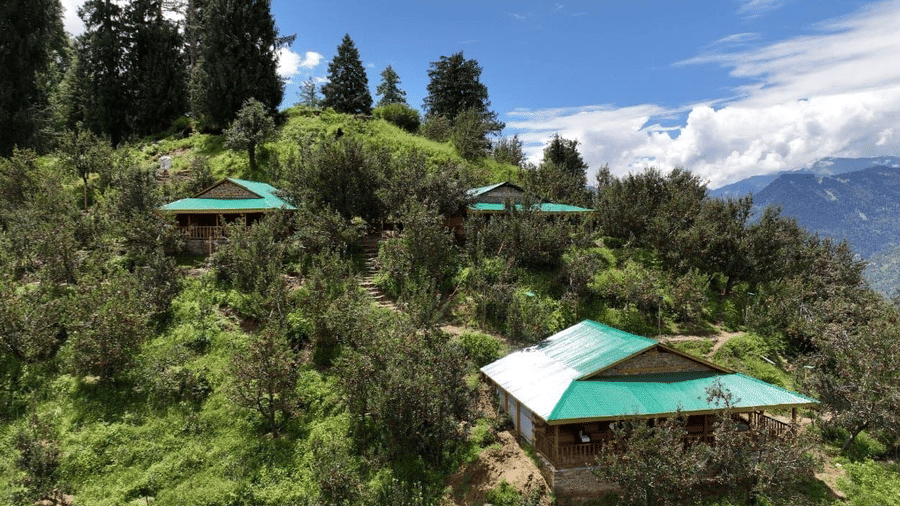 A beautiful landscape featuring 3 cottages amid lush green vegetation at Amara Upepo - The Sky Village, Manali, with a clear blue sky in the background.
