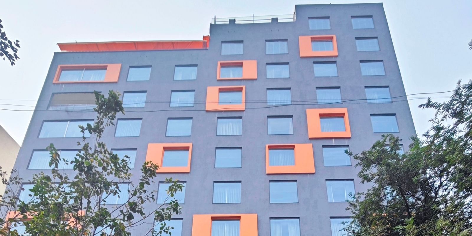 A modern, multi-story hotel building with a dark grey facade and bright orange window accents. The hotel has a covered parking area in front, and there are trees and shrubs surrounding the building.