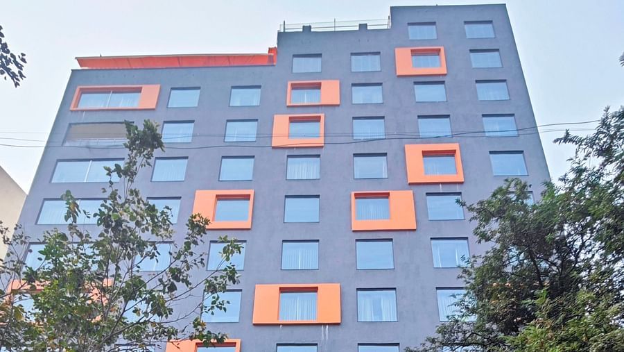 A modern, multi-story hotel building with a dark grey facade and bright orange window accents. The hotel has a covered parking area in front, and there are trees and shrubs surrounding the building.