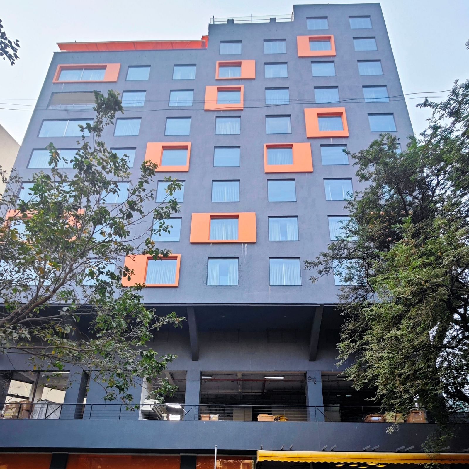 A modern, multi-story hotel building with a dark grey facade and bright orange window accents. The hotel has a covered parking area in front, and there are trees and shrubs surrounding the building.