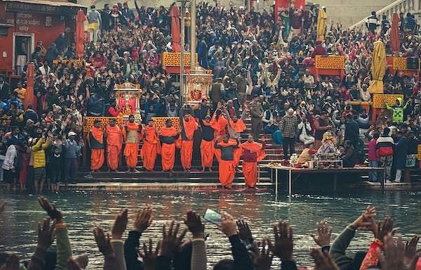 A large gathering by the Ganges River bank creates a vibrant scene filled with people engaged in various activities, from worship and meditation to socializing. The river’s flowing waters and surrounding landscape enhance the sense of community and spiritu
