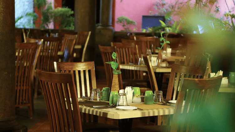 The dimly lit, empty tables and wooden chairs in what appears to be a restaurant at Grasshopper reataurant Uday Samudra Leisure Beach Hotel.