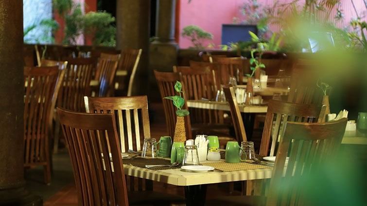 The dimly lit, empty tables and wooden chairs in what appears to be a restaurant at Grasshopper reataurant Uday Samudra Leisure Beach Hotel.