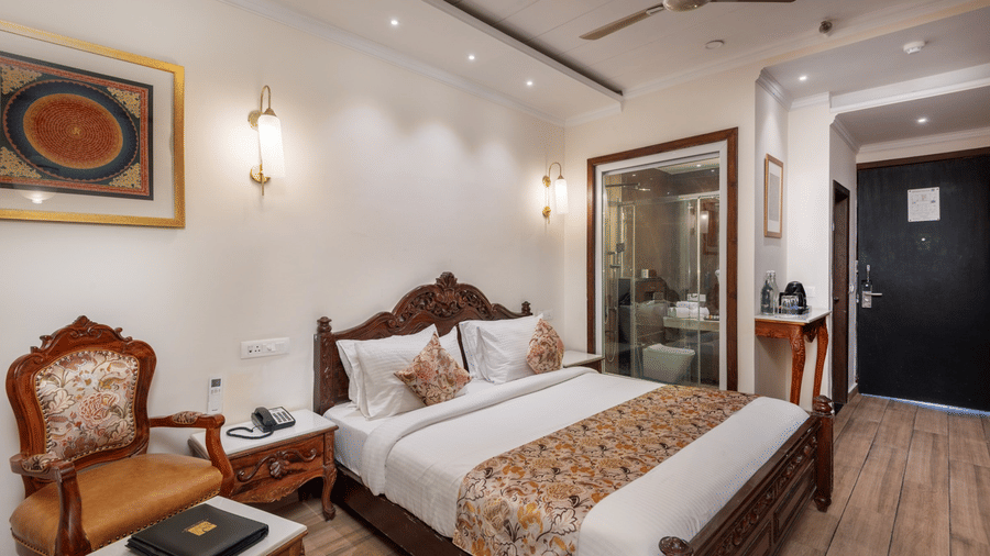 Hotel room at Quality Inn Dharamshala with a carved wooden king bed, floral bedspread, vintage armchair, wall sconce, and glass bathroom partition visible in the background.