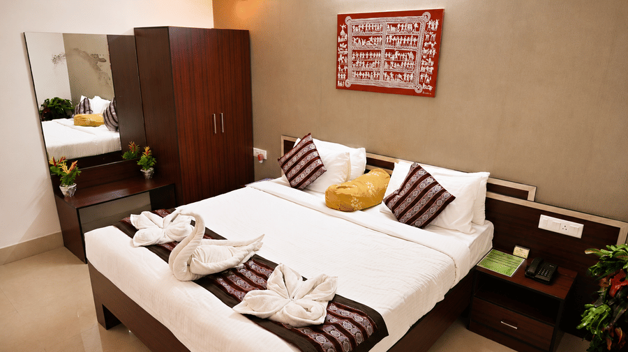 Hotel room with a bed dressed in white linen, swan-shaped towel art, patterned cushions, a wooden wardrobe and a wall-mounted mirror nearby.