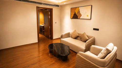 A suite features a fabric sofa, an armchair, and a black marble coffee table on wooden flooring near an open doorway at Pride Plaza Indore.