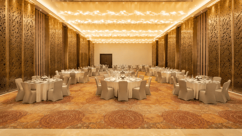 Round dining tables with chairs are arranged inside a hall featuring golden patterned walls, a patterned carpet, and lights at Pride Plaza Indore.