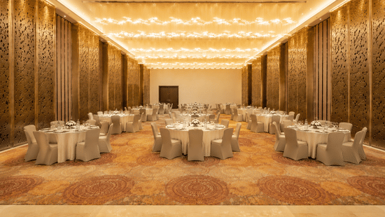 Round dining tables with chairs are arranged inside a hall featuring golden patterned walls, a patterned carpet, and lights at Pride Plaza Indore.