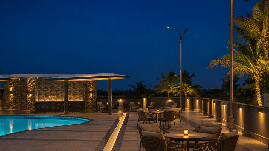 Poolside deck at Pride Elite Mandarmani at dusk, with a lit swimming pool, stone-clad bar counter, lounge seating around tables with candles, and palm trees.