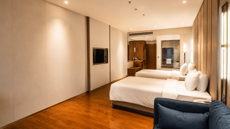A Superior Twin Room at Pride Plaza Indore featuring two single beds, a wall-mounted television, and an upholstered sofa in the foreground.