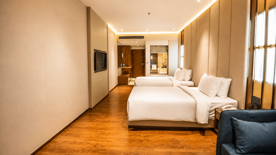 View of a Superior Twin Room at Pride Plaza Indore showing 2 single beds, built-in wall lighting, and a partial view of an armchair.