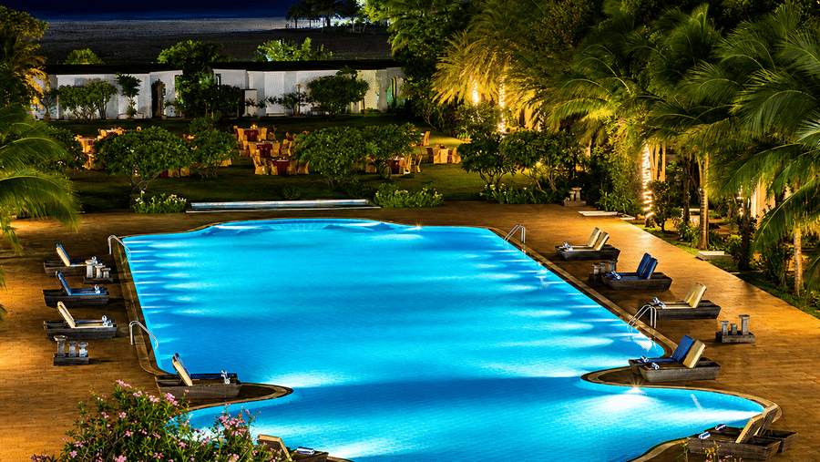 Swimming pool beautifully illuminated at night with lounge chairs and palm trees at Chariot Beach Resort, offering a magical evening setting.