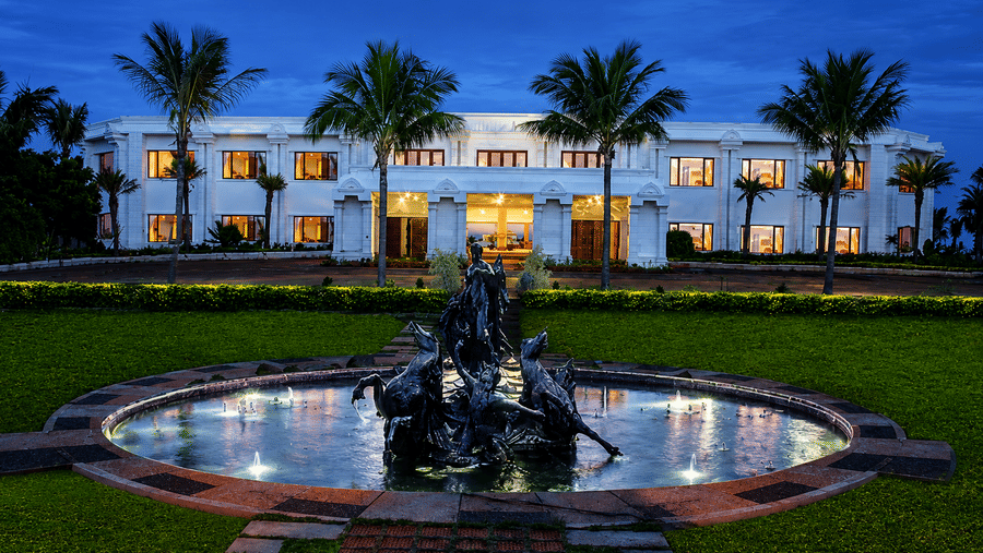 Outside view of Chariot Beach Resort showcasing an illuminated facade, elegant fountain, and palm trees against the evening sky.