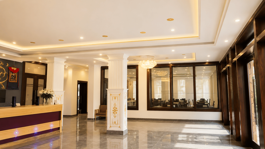 Hotel lobby with a reception desk, marble pillars, glass-panelled offices and polished tiled flooring with warm ceiling lighting throughout.