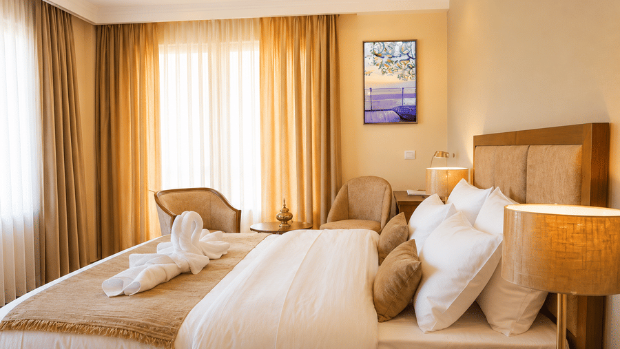 Hotel room with a bed dressed in white linen, swan-shaped towel art, gold cushions, a wall-mounted artwork and long golden curtains nearby.