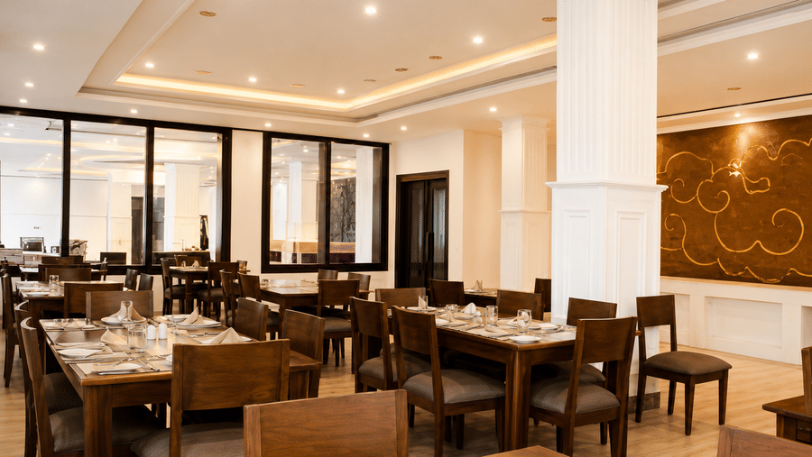 Restaurant dining area with wooden tables and chairs, decorative pillars, patterned wall panels and recessed ceiling lighting throughout it.