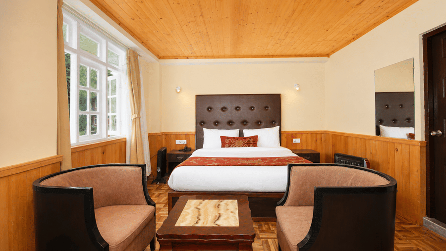 Deluxe Room featuring a comfortable seating area with two armchairs and a small table sits at the foot of a bed. This interior at Orsino Khangri Karpo Retreat & Spa, Lachung features extensive wooden panelling.