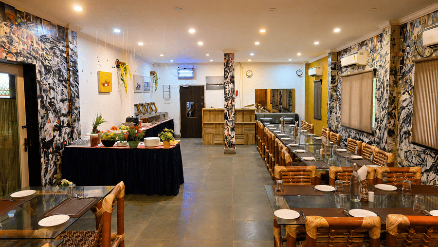 Restaurant seating area with wooden cane chairs, dark wooden tables, patterned wall art and a buffet counter set out in the background nearby.
