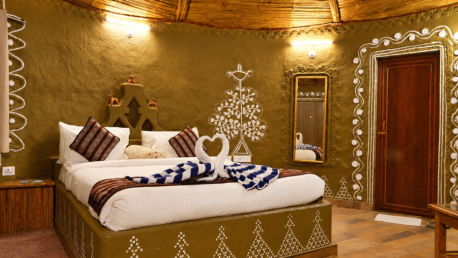 Tribal cottage bedroom with hand-painted tribal wall art, a bed dressed in white and blue linen, a wooden door and a thatched ceiling above.