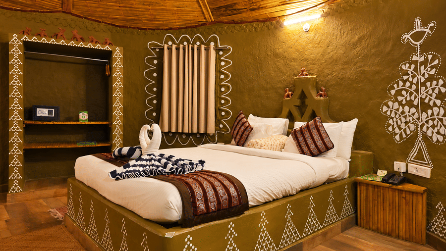 Tribal cottage bedroom with hand-painted tribal wall art, a bed dressed in patterned linen, a thatched ceiling and a curved mud wall design.