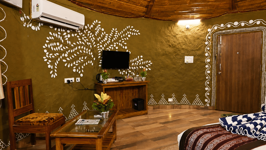 Tribal cottage seating area with a wooden coffee table, a chair, hand-painted tribal wall art, a wall-mounted television and a thatched ceiling.
