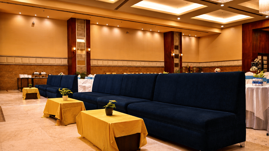 Banquet hall lounge area with navy blue sofas, gold side tables, decorative pillars and warm ambient ceiling lighting spread across the hall.