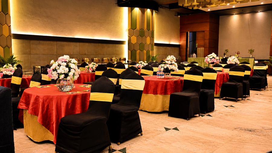 Banquet hall with round tables dressed in red and black linen with gold accents, floral centerpieces and warm ambient lighting spread overhead.