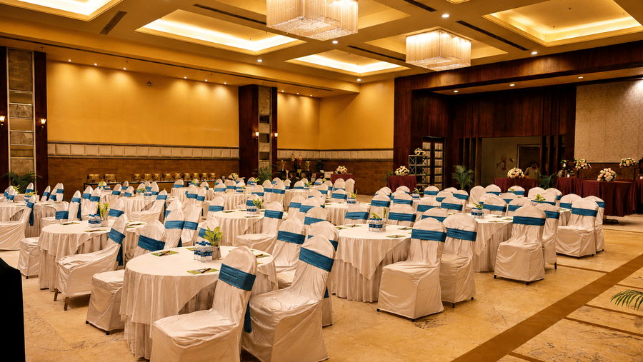 Banquet hall set up theatre-style with rows of white chair covers and teal sashes, chandeliers overhead and warm gold ambient lighting round.