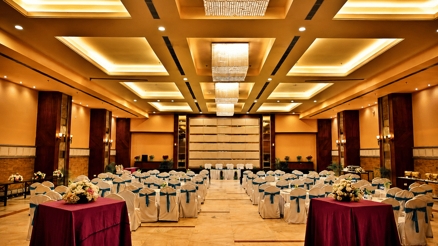 Banquet hall wide view with rows of round tables in white and maroon linen, a coffered ceiling and warm gold lighting spread throughout the hall.