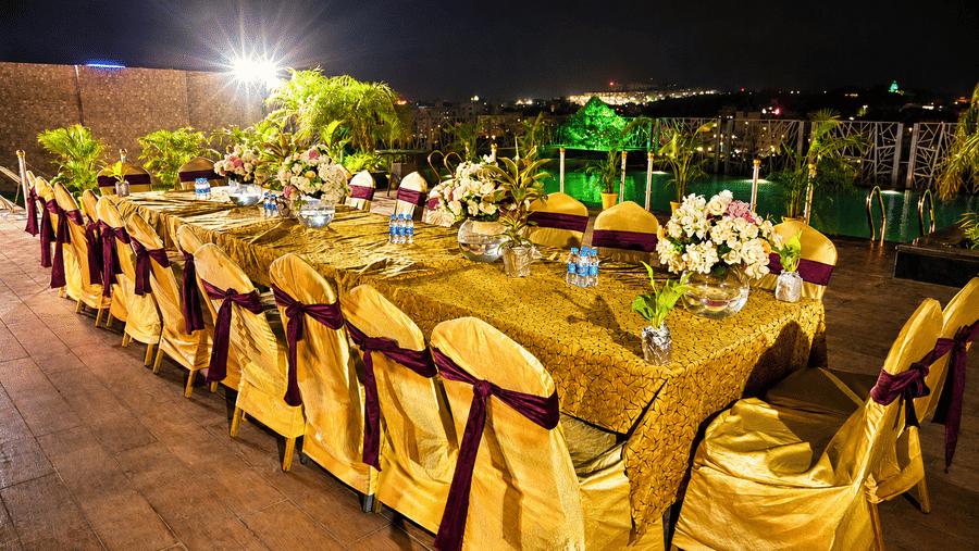 Outdoor poolside deck set up at night with a long banquet table, gold chair covers, floral centrepieces and string lights across the venue.