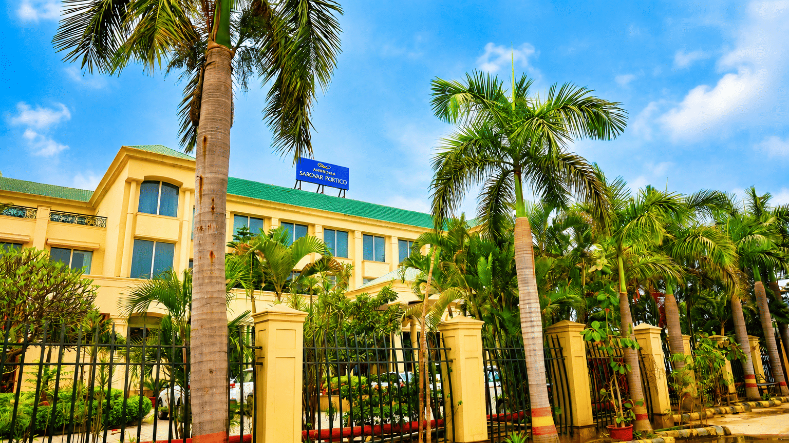 Front facade of Ambrosia Sarovar Portico, Haridwar, with a palm tress lined entrance and boundary gate view.