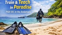Scuba diver in full gear standing on sandy beach with fins in foreground, turquoise water and green cliffs behind, text promoting PADI IDC training in the Andamans.