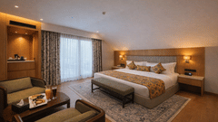 A spacious bedroom at Mountnoor Sarovar Premiere featuring a king size bed, a seating area with 2 chairs, and a large window with heavy drapes.